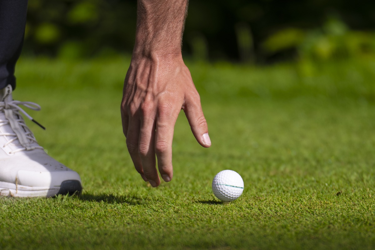 Detail: Hand legt den Golfball ins Gras, daneben der Golfschuh