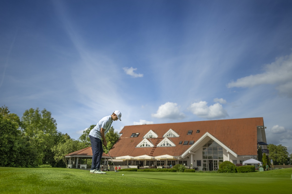 Spieler beim Schlag auf dem Fairway vor dem Clubhaus, darüber weiter Sommerhimmel