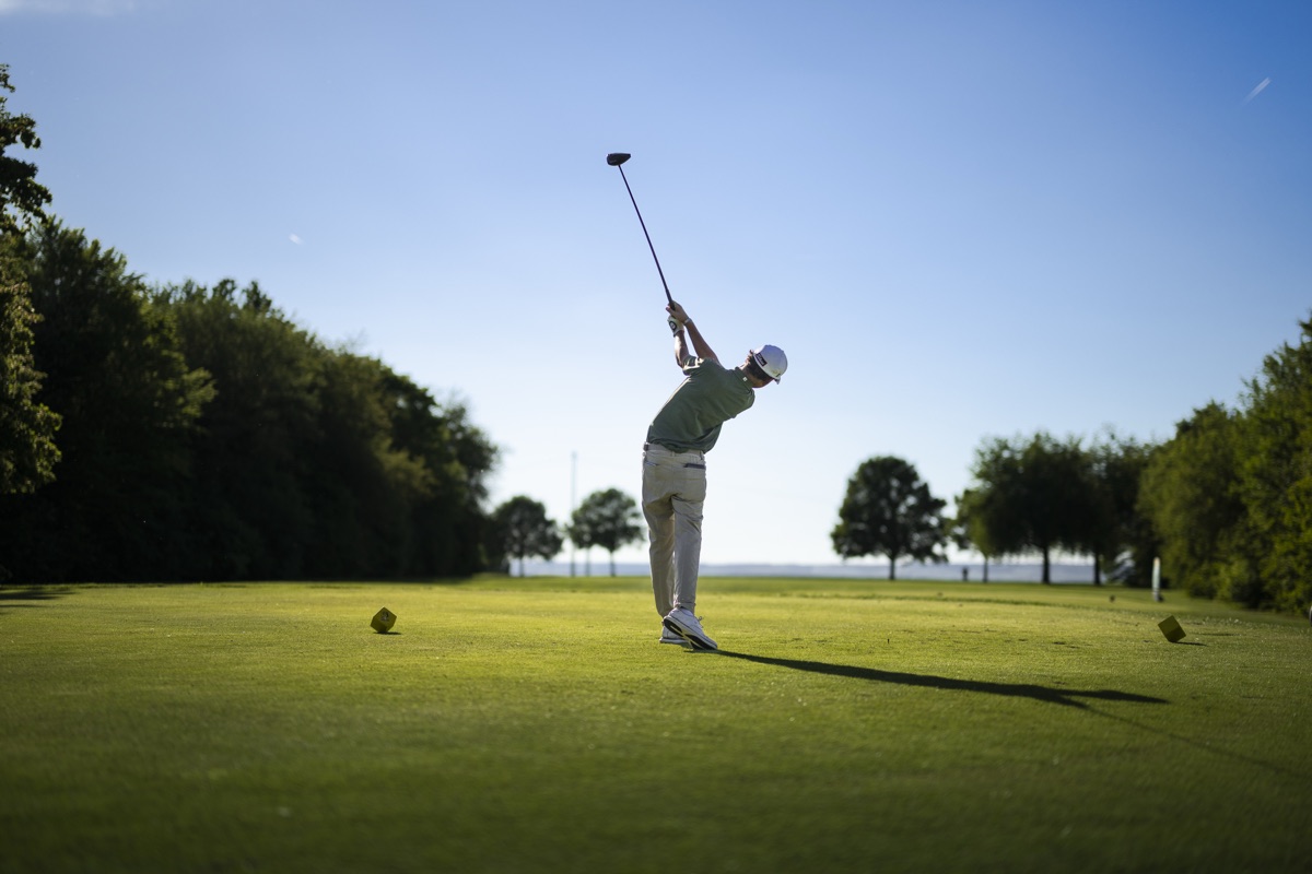 Abschlag: Spieler im Durchschwung auf dem Tee, Parklandschaft und blauer Himmel